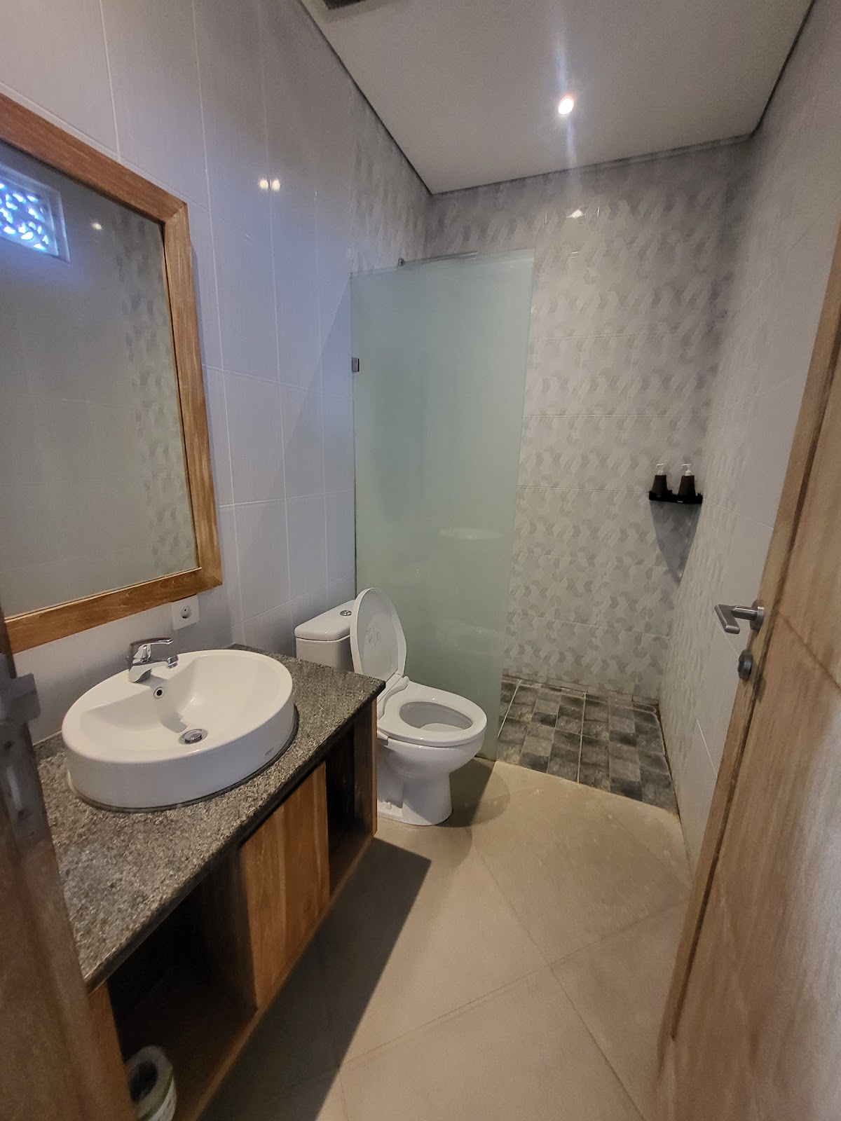 Modern bathroom with round white sink, granite countertop, wooden framed mirror, toilet, and walk-in shower with glass partition. Clean white tile walls and well-lit with recessed ceiling lights.